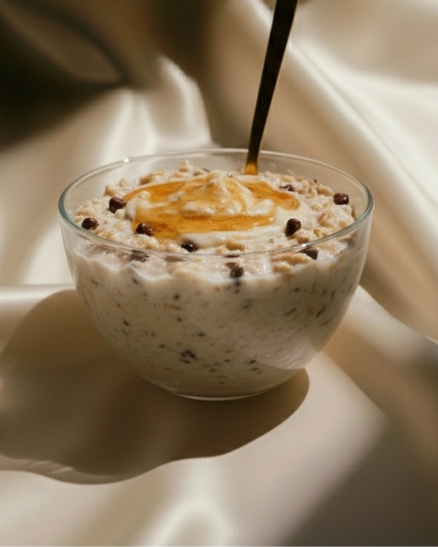 Glass bowl of overnight oats with chocolate chips, cream and honey being drizzled from a gold spoon