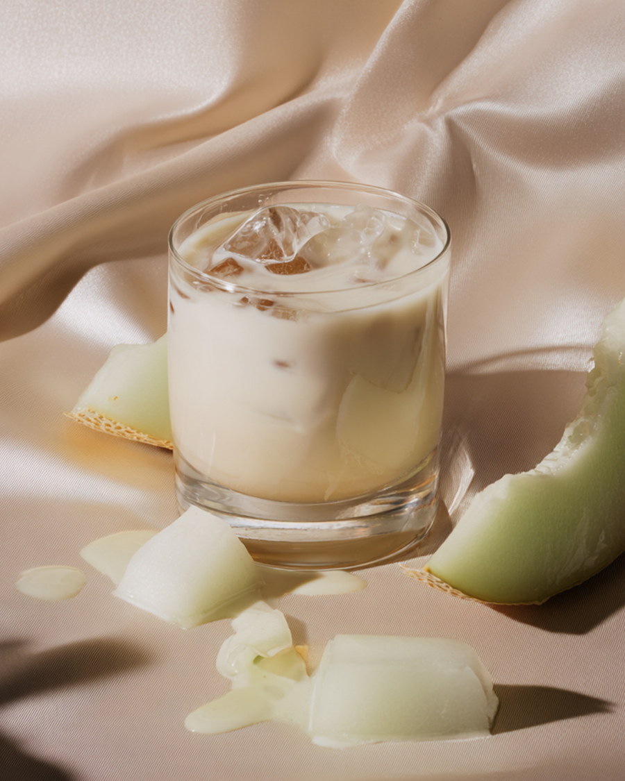 Iced honeydew melon collagen milk in a heavy tumbler, creamy and pale over ice, with honeydew wedges and cubes on the table