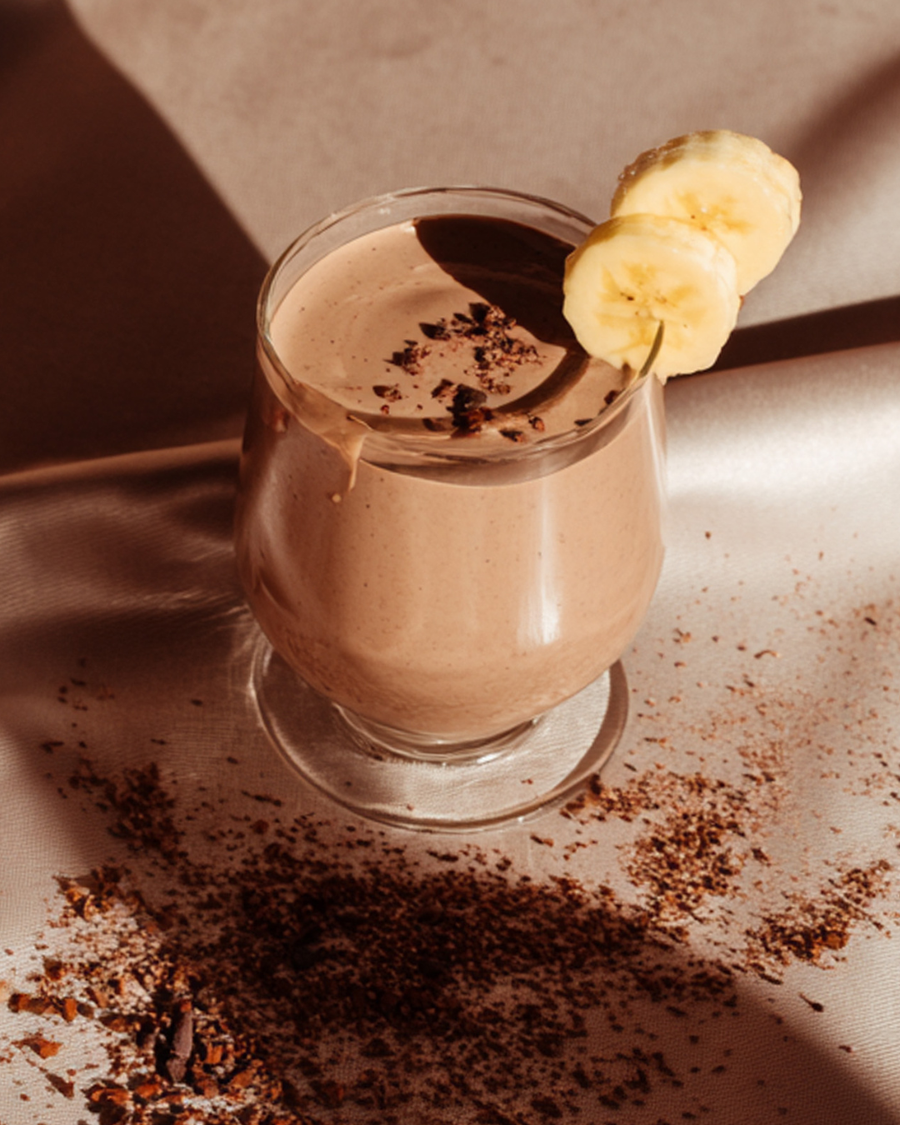 Blended chocolate banana collagen smoothie in a stemmed glass, topped with cacao nibs and two banana slices on the rim