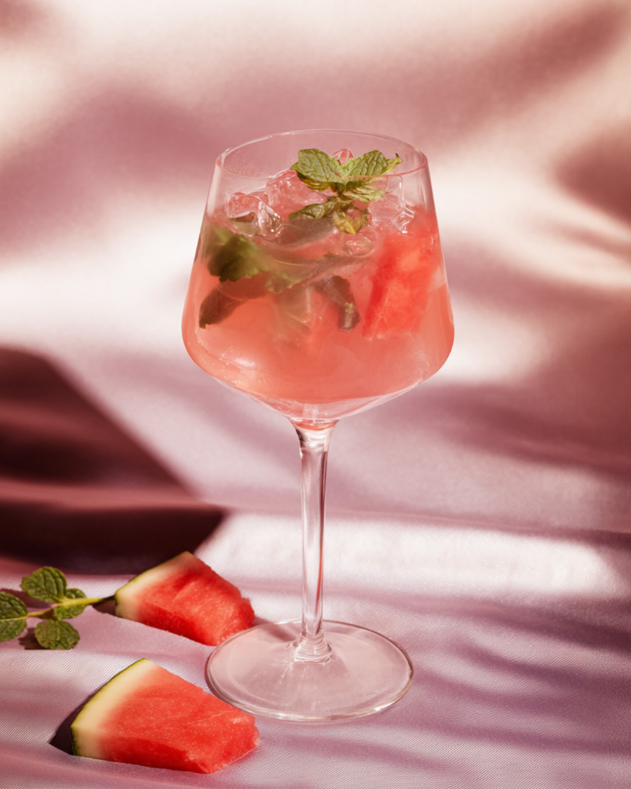 Pink watermelon collagen spritz in a stemmed wine glass over crushed ice with mint, watermelon wedges alongside