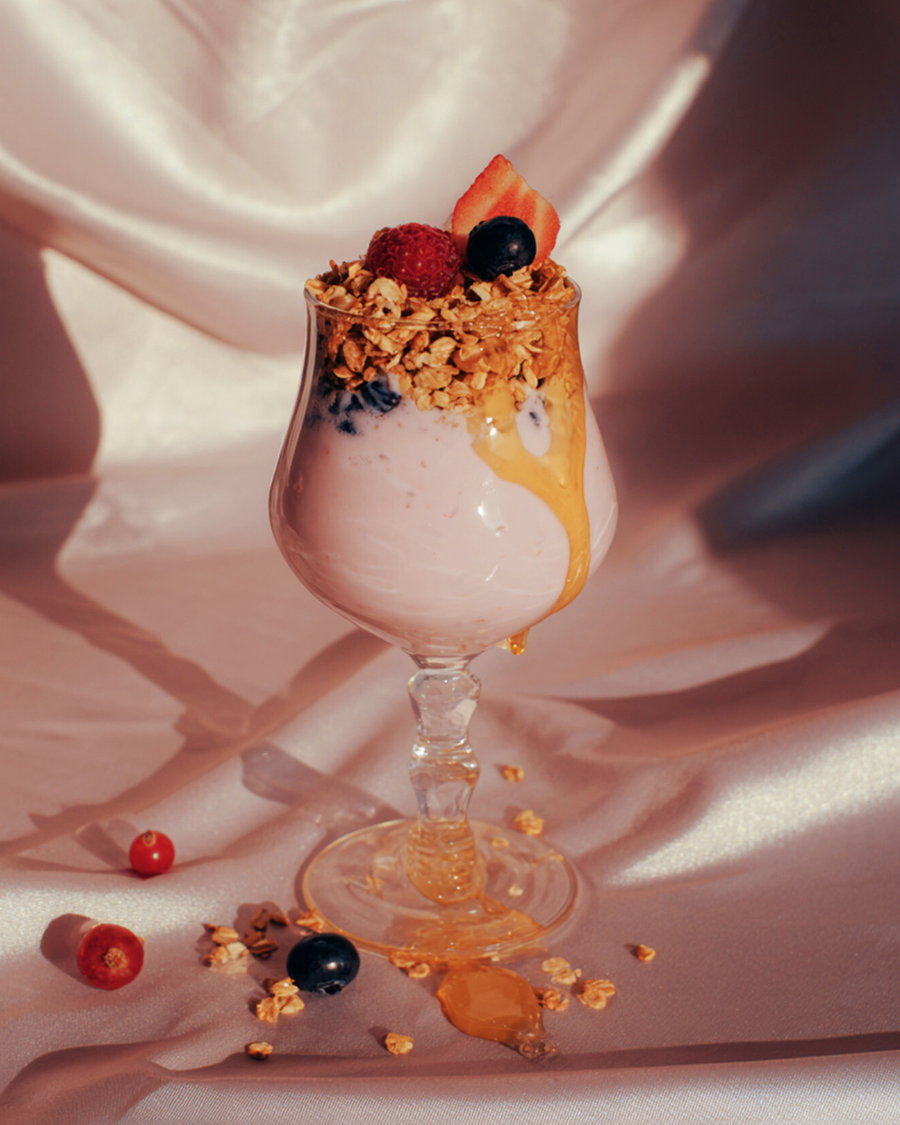 Layered collagen yoghurt parfait in a stemmed glass with granola, blueberries, raspberry and honey running down the side